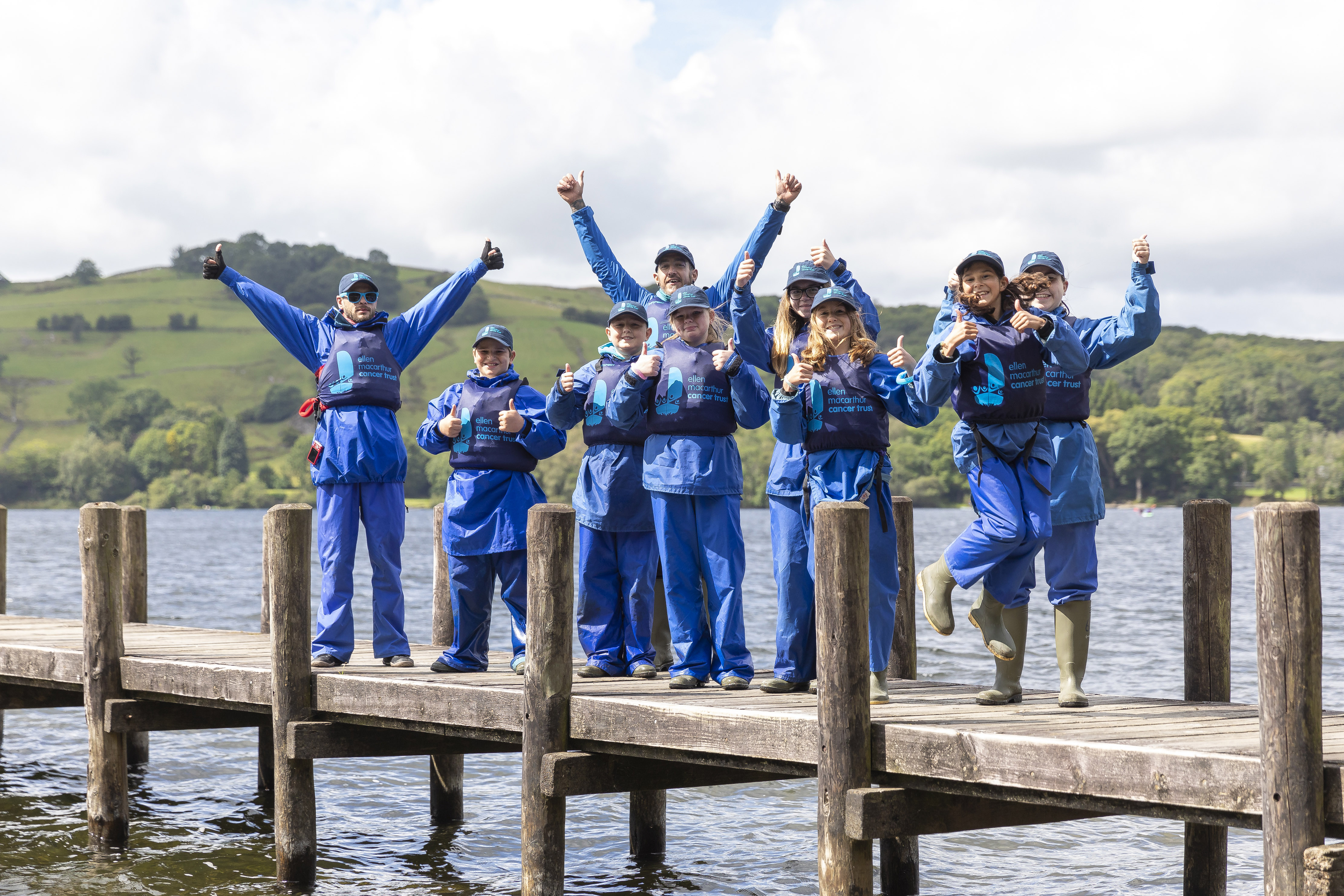A group of young people and volunteers wearing Trust branded waterproof clothing are smiling and jumping by a lake
