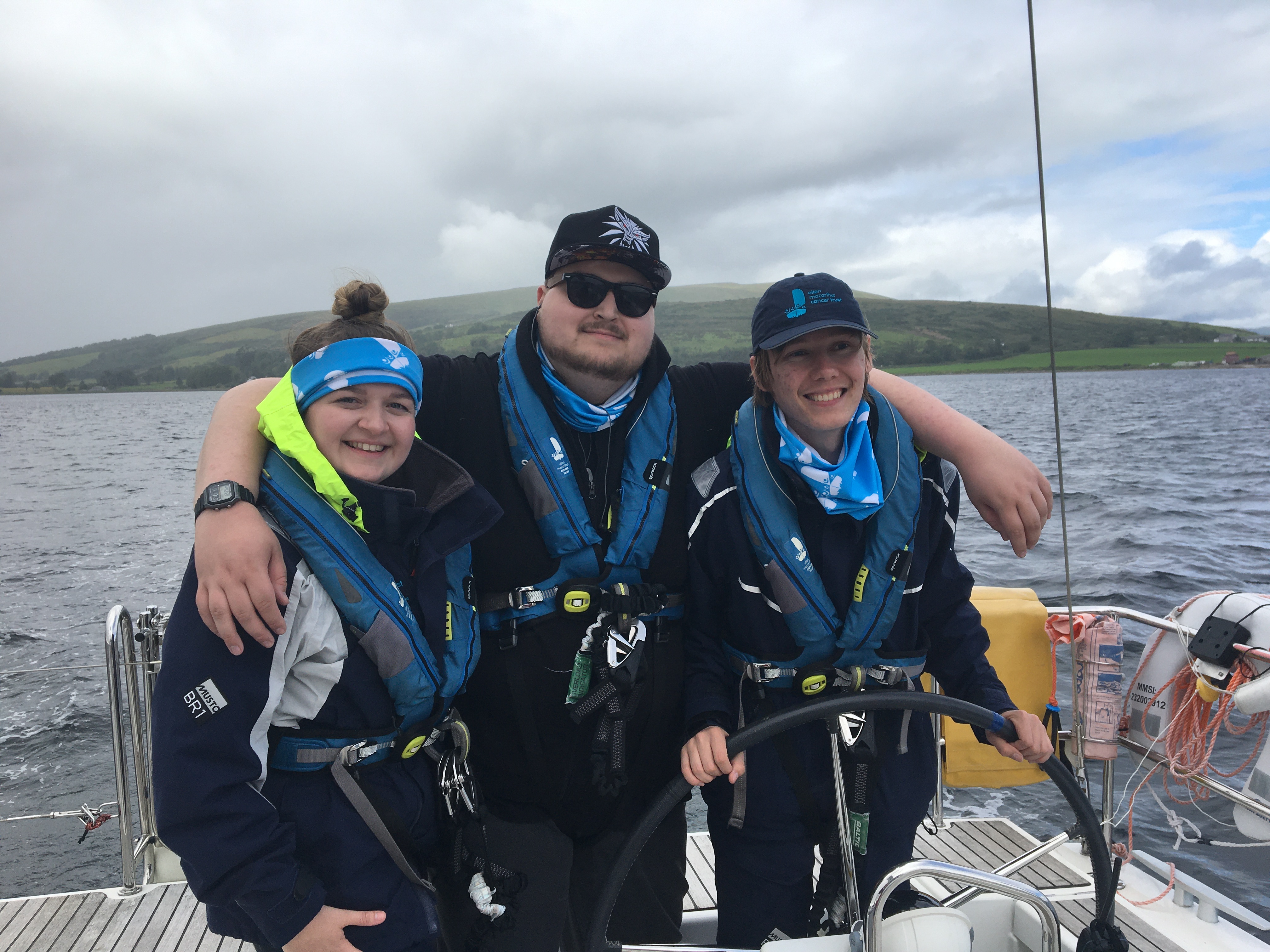 Lucy smiling at the camera with her arm round two crew mates while sailing on a Trust trip in Largs, with grey waters and green hills behind her