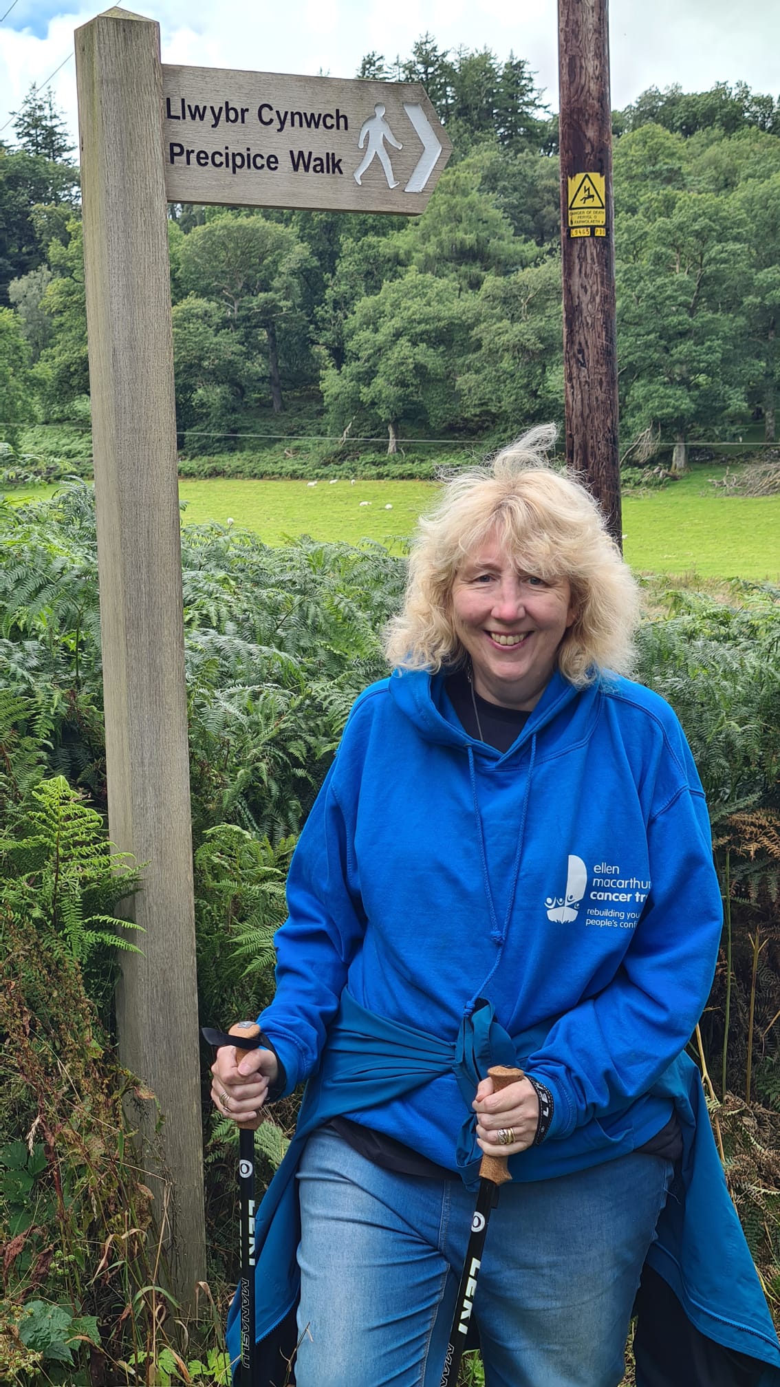 Katharine on her walk in Wales, smiling at the camera while wearing a blue Ellen MacArthur Cancer Trust hoodie, standing beside a sign that says Precipice Walk in English and Welsh