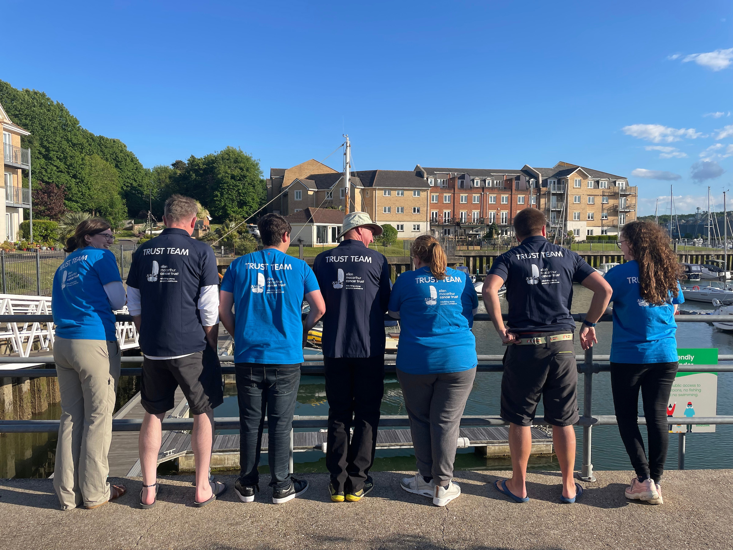 Volunteer and skippers with their backs to the camera, showing their t-shirts which say 'Trust team' along the back, as they look out over the sunny East Cowes marina