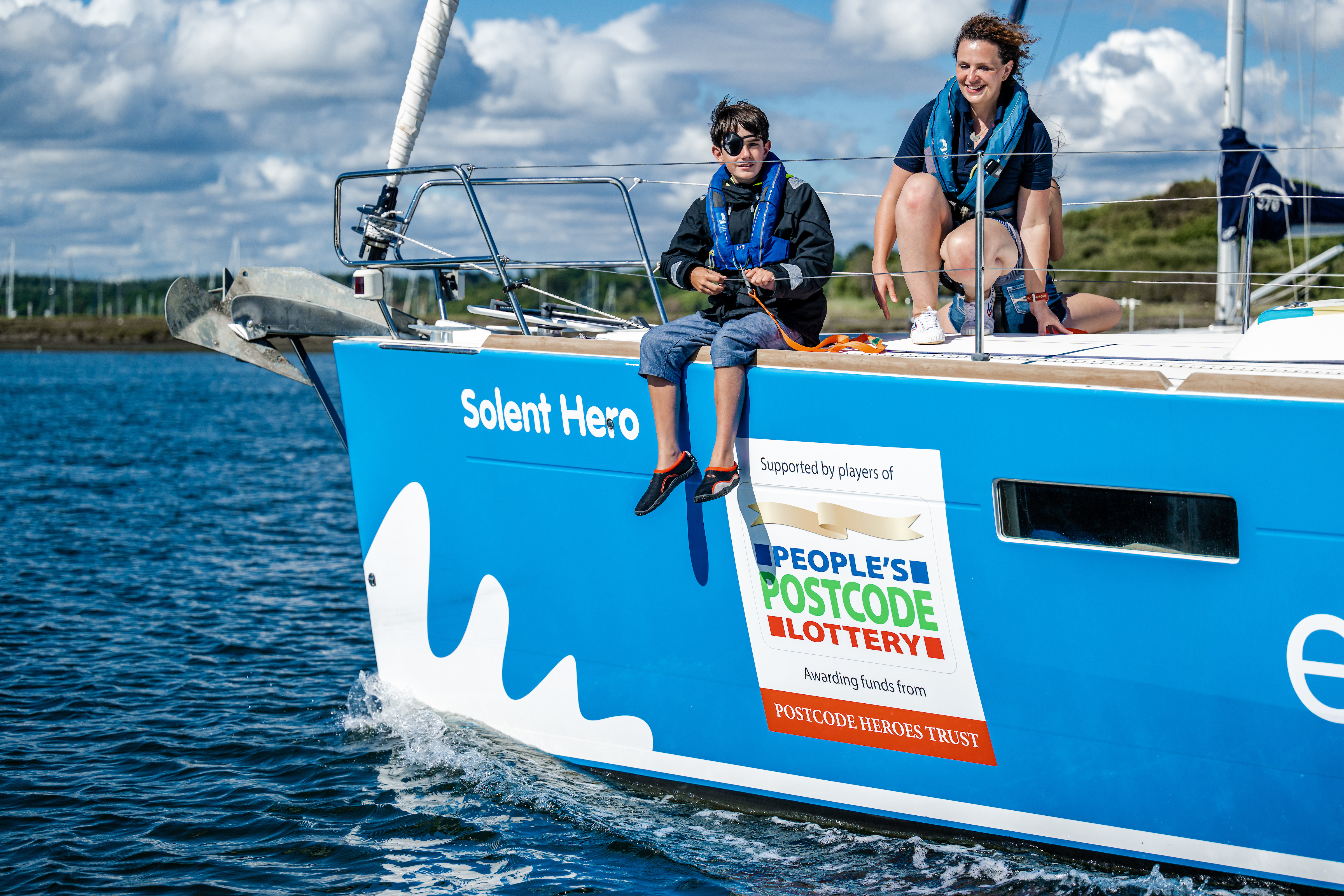 The Trust's yacht Solent Hero on a sunny day with a young person sitting with their legs over the side, with a focus on the People's Postcode Lottery on the bow