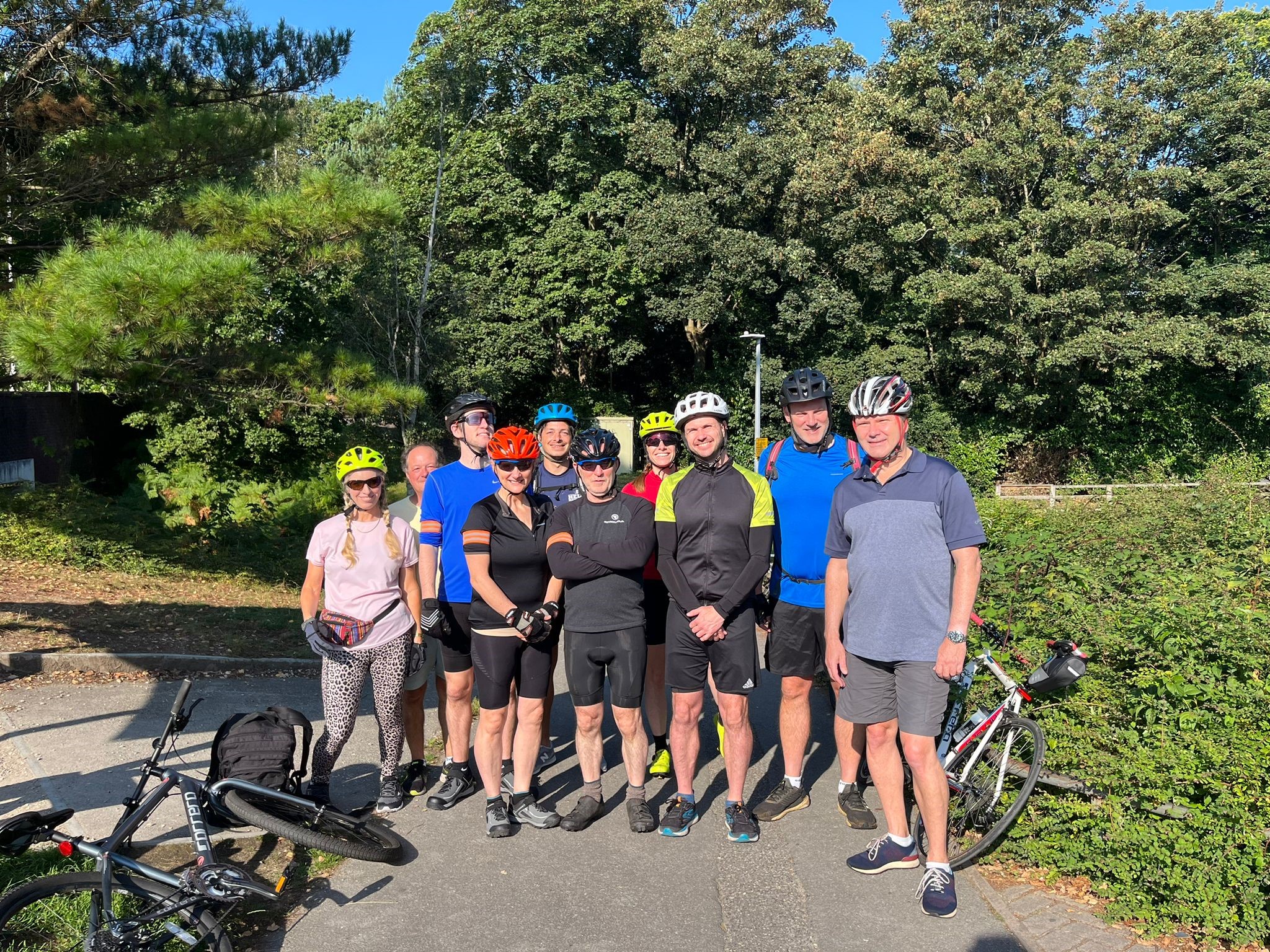 Tim and his group of cycling colleagues standing on a cycle path on a sunny day, smiling at the camera while dressed in cycling gear