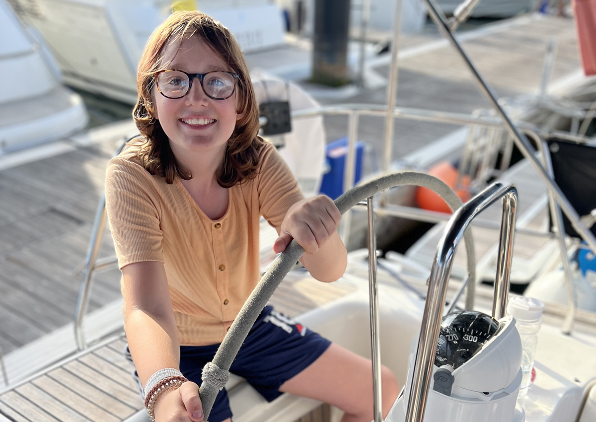 Yasmin sitting at the helm of the yacht while docked in a marina, smiling at the camera 