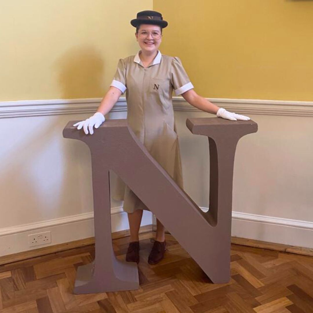 Lucy pictured in the famous Norland's nanny uniform of brown dress, top hat and white gloves posing next to the Norland Nanny logo.