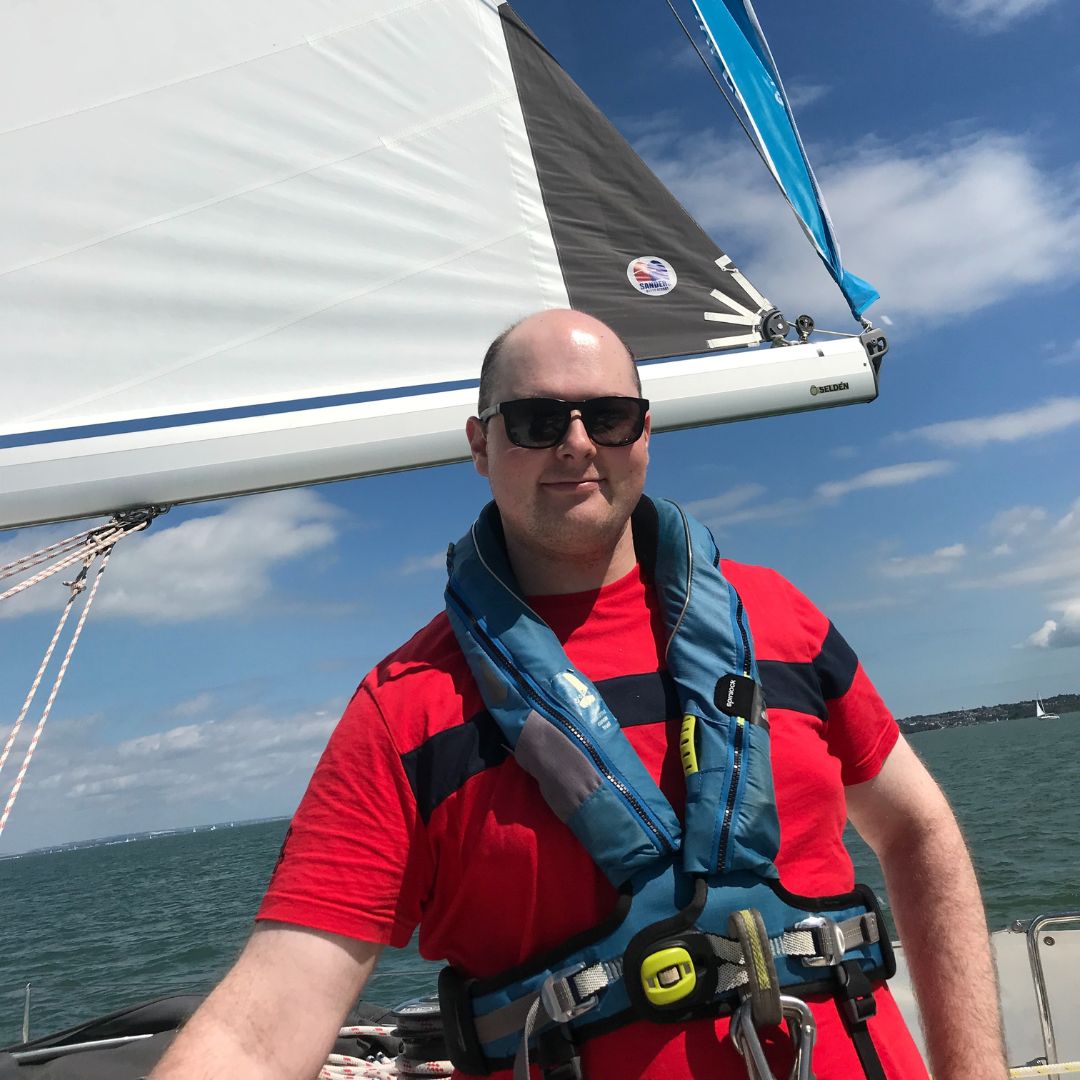 Martin smiling on one of the Trust yachts wearing a red t-shirt and blue life jacket with the sea in the background on a beautiful sunny day.