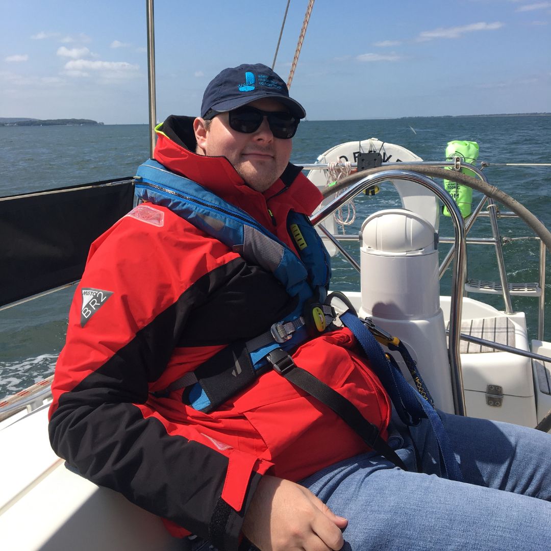 Martin sat chilling on one of the Trust yachts wearing a red t-shirt and blue life jacket with the sea in the background on a beautiful sunny day.